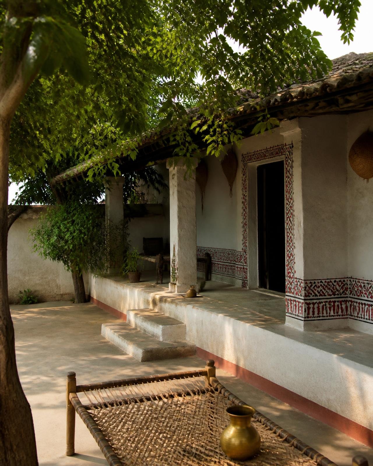 A shaded Mithila village courtyard with painted folk borders, a charpai and brass vessel